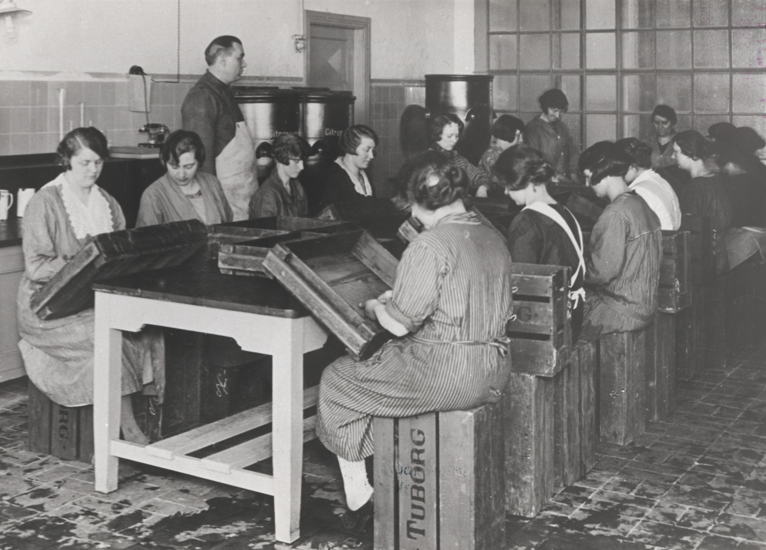 Kvinder sidder omkring et bord på bryggeriet Tuborg. De sidder på ølkasser og arbejder med noget, vi ikke kan se ved bordet. En mand står og holder opsyn med dem. Billedet er i sort hvidt fra 1920. 