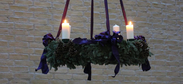 Adventskransen af gran og med lilla bånd hænger i Stavnsholtkirken. To lys er tændt og man kan se den rå murstensmur i baggrunden.
