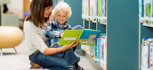 An kvinde i hvid bluse og med mørkt hår, sidder på gulvet foran en reol på et bibliotek med en lyshåret barn på skødet og læser højt fra en bog.