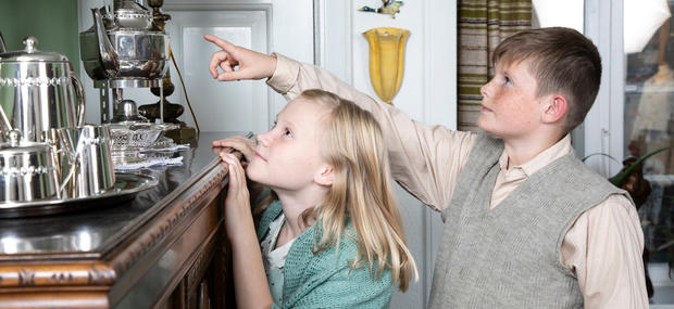 To børn - en dreng og en pige - i 1930'er stuen. De kigger begge op på et billede, mens drengen peger på det.