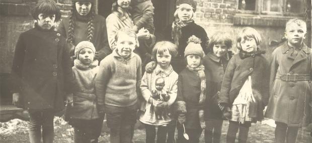 En gruppe børn fotograferet i en baggård. Børnene står på to rækker og kigger på fotgrafen, en af dem har en ske i hånden, en anden en dukke. Fotoet er sort hvidt. 