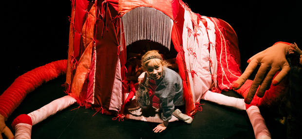 Et billede af 24 hemmeligheder på Teater ZeBU. En lille pige kraver ud af en hule. Hulen er en figur med lange arme og ben, som er syet af stoffer i røde nuancer.