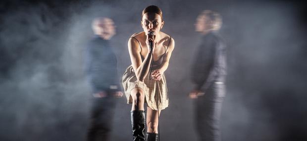 Et billede af Antigone på Teater Zebu. I forgrunden står en kvinde foroverbøjet med en mikrofon i hånden. I baggrunden er der røg, og man ser to udviskede mænd stående på hver siden side af kvinden. De kigger på hinanden.