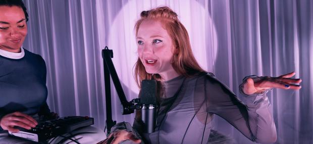 Et billede af Natten er lavet af glas på Teater ZeBU: En ung kvinde med rødt hår taler ind i en podcast-mikrofon i midten af billedet. Til venstre ses en ung kvinde med hørebøffer på, der kigge smilende ned på en lydboks. Baggrunden er ét stort lyselilla gardin.