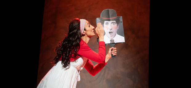 Et billede af Vai Via på Teater Zebu. En kvinde med langt, mørkt og krøllet hår står med siden til og hvisker et skilt i øret, som hun holder. Skiltet portrætterer et mandeansigt med en hat på, som laver store øjne og åben mund.