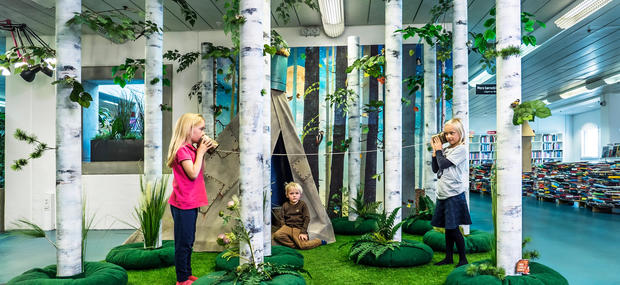 Billedet viser en kreativ indendørs installation, der forestiller en skov med høje birketræer og grønt græs på gulvet. I midten af installationen er et lille telt, og tre børn leger i skoven. To af børnene står på hver sin side af installationen og holder papkopper forbundet med en snor op til deres øre og mund, som om de kommunikerer gennem dem. Det tredje barn sidder på græsset foran teltet og kigger fremad. I baggrunden er der reoler fyldt med bøger,hvilket indikerer, at scenen finder sted i et bibliotek