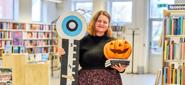 bibliotekaren står med en stakbøger, et Jack-o-Lantern og et skræmmometer