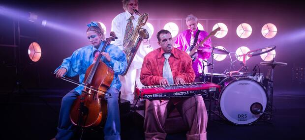 Fire mennesker i pastelfarvede alt for store jakkesæt i hvid, lysblå, aprikos og lysreød, spiller på saxofon, cello, keyboard og guitar med store lamper bagved sig.