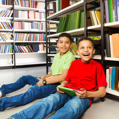 To drenge i henholdsvis rød og grøn t-shirt og begge med cowboybukser sidder på gulvet med hver sin bog, lænet op af en bogreol på biblioteket.