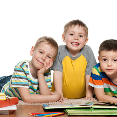 Tre drenge i børnehaveklassealderen ligger ovenpå en stabelbøger og ser glade ud mod os