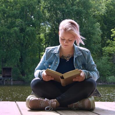 Blond ung kvinde i cowboy jakke, sidder i skrædderstilling i solen og læser i en bog på en træterrasse udenfor.